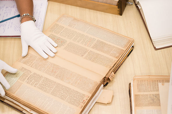 Close-up of two hands wearing white gloves holding up pages in an aged book laying flat on a light wood table, with other aged books laying flat open on the same table