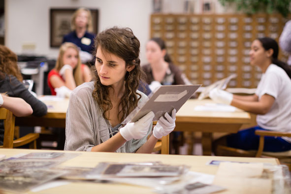 A seated young woman looking down at plastic-covered photographs on laying on a light wood table, while holding up a protected photograph wearing white gloves