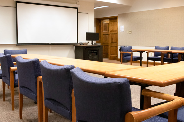 light brown tables, with blue chairs tucked underneath, facing a wall with a dry erase board with a projector screen in front, and a black podium with a black desktop 