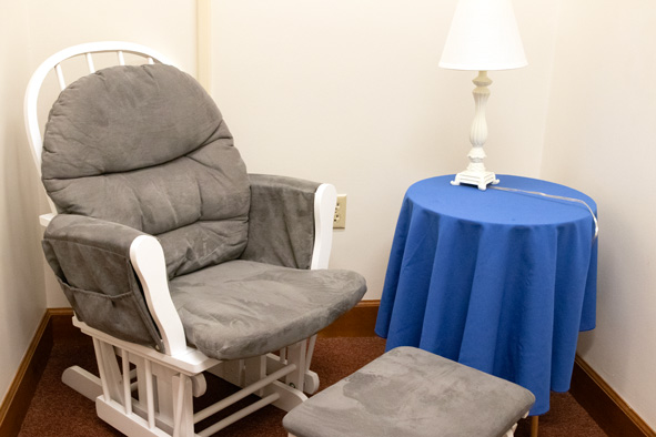 a white rocking chair with gray velvet cushions and matching foot rest next to a small round end table, covered with a blue tablecloth, and a white lamp sitting on top