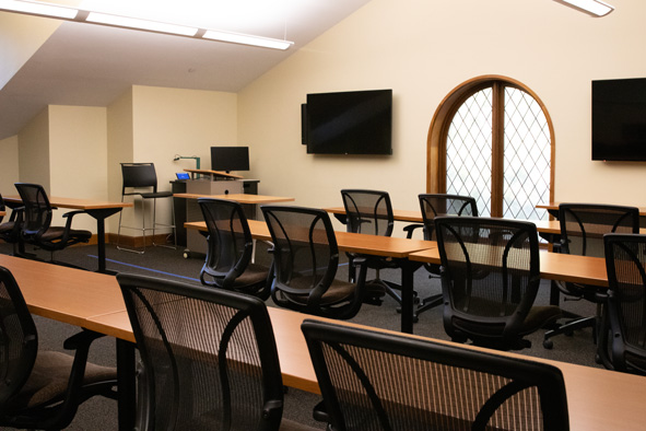 Brown tables with black chairs facing a wall with an arched window. On each side of the window are two black television screens.